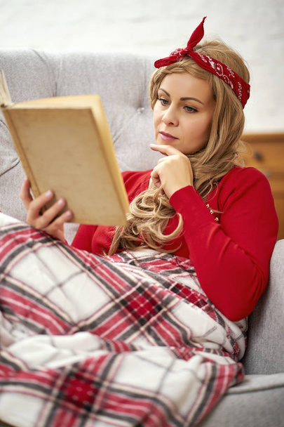 νέοι χαμογελώντας όμορφη γυναίκα που κάθεται και διαβάζοντας ένα βιβλίο, τυλιγμένο σε μια κουβέρτα, κατά την περίοδο των Χριστουγέννων - Φωτογραφία, εικόνα