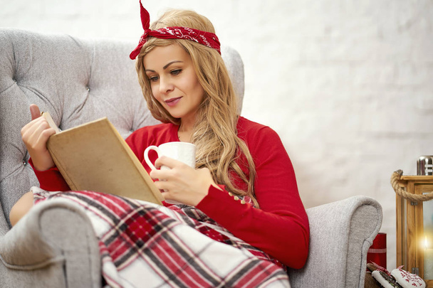 young beautiful woman reading a book in an armchair with blanket and tea during Christmas time - Photo, Image