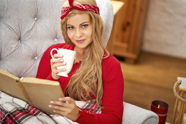 Молодая красивая женщина читает книгу в кресле с одеялом и чаем во время Рождества
 - Фото, изображение