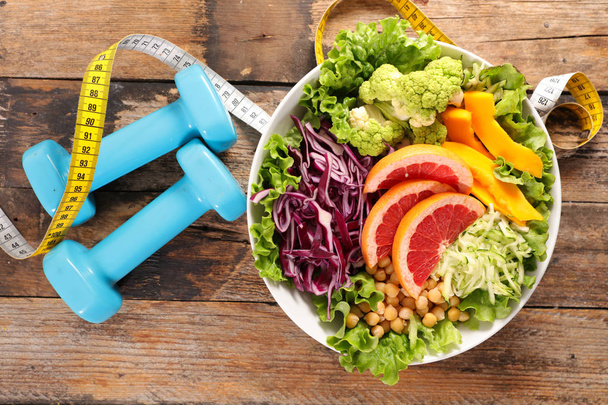 Healthy salad with dumbbells and measuring tape on wooden table, diet food concept - 写真・画像