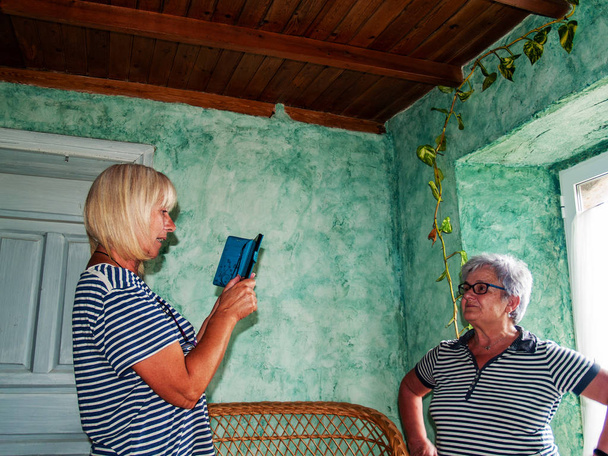 Kaksi aikuista naista hymyilee ja ottaa kuvia älypuhelimella maalaistalossa
 - Valokuva, kuva
