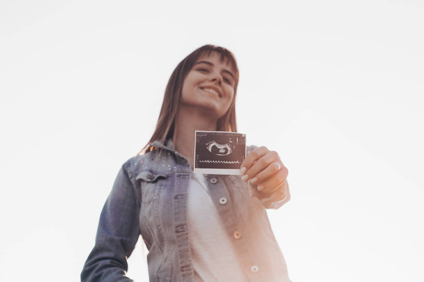 Młoda kobieta w ciąży trzyma zdjęcie USG o zachodzie słońca i ogarnięcie jej brzuch. 4 miesiąca ciąży. Koncepcja macierzyństwo. Stonowanych zdjęcie - Zdjęcie, obraz