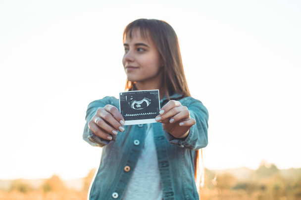 Młoda kobieta w ciąży trzyma zdjęcie USG o zachodzie słońca i ogarnięcie jej brzuch. 4 miesiąca ciąży. Koncepcja macierzyństwo. Stonowanych zdjęcie - Zdjęcie, obraz