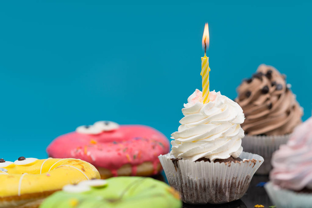 Bolo de festa com gelo cremoso girado e uma única vela de aniversário em chamas entre bolos variados sobre azul no espaço de cópia
 - Foto, Imagem