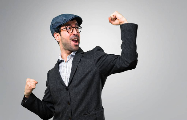 Handsome modern man with beret and glasses celebrating a victory on grey background - 写真・画像