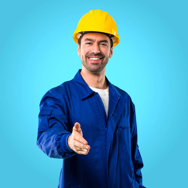 Young workman with helmet shaking hands for closing a good deal on blue background - Photo, Image