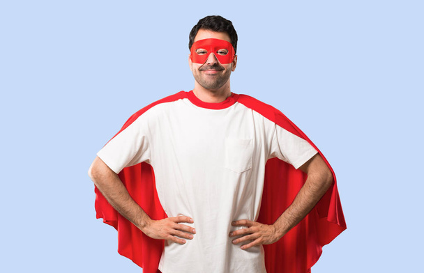 Superhrdina muž s maskou a červená mys pózuje s rukama v kyčli a směje se, při pohledu na frontu na izolované modré pozadí - Fotografie, Obrázek