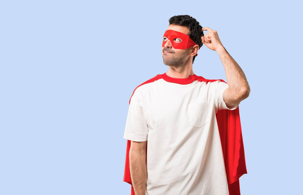Superhero man with mask and red cape having doubts and with confuse face expression while scratching head on isolated blue background - Foto, afbeelding
