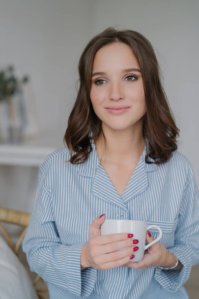 Indoor shot of dreamy young European woman with charming look, dressed in striped pyjamas, drinks coffee, thinks about something, enjoys calm domestic atmosphere. People, drinking and home concept - Photo, Image