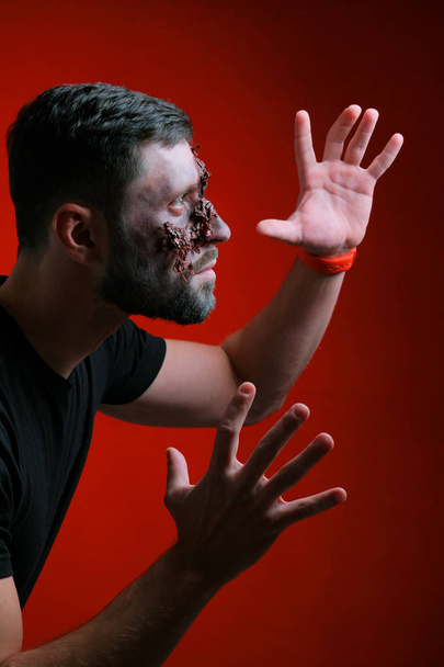 The guy in profile with Halloween-style makeup raised his hands and spread his fingers. Close-up. A young man with a torn wound and bruises on his face. Dark red background. - Foto, immagini