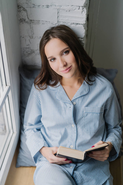 Beautiful relaxed woman with carefree expression, dressed in nightclothes, reads book on window sill in morning, enjoys spare time, likes her hobby. Vertical shot of teenage girl with literature - Foto, afbeelding