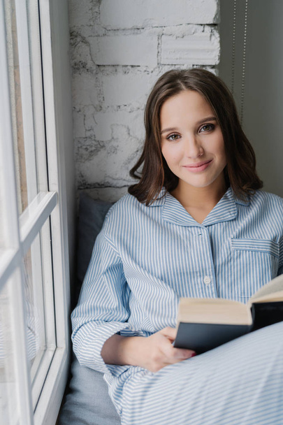 Photo of beautiful woman dressed in casual clothes, wears pyjamas, sits on window sill, reads romantic story in book, enjoys coziness and comfort. Young female with textbook at home. Leisure concept - Foto, Bild