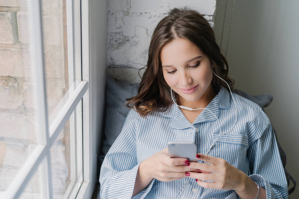 Charming woman enjoys romantic playlist, perfect sound in earphones, holds modern smart phone, dials number of boyfriend, sits on window sill, connected to wireless internet. Rest and home concept - 写真・画像