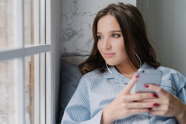 Thoughtful brunette woman listens audio book, holds modern smart phone in hands, enjoys perfect sound in earphones looks pensively at window dressed in casual clothes. Female listens music in playlist - Foto, imagen
