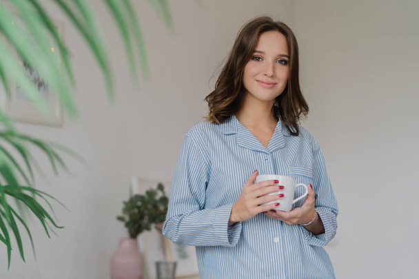 Waist up shot of pleasant looking young woman dressed in striped pyjamas, drinks hot beverage after awakening, has sparre time at home, stands indoor against cozy interior. People and rest concept - Fotó, kép