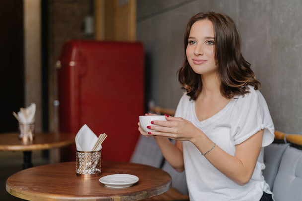 Довольно беззаботная молодая женщина в повседневной белой футболке, имеет счастливое лицо, пьет ароматный кофе, проводит свободное время в ресторане, сидит за круглым деревянным столом, имеет вдумчивое выражение на расстоянии
. - Фото, изображение