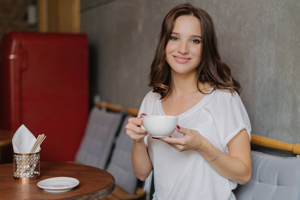 Smiling woman with tender smile, holds cup of tea or coffee, enjoys good rest, poses in cozy cafe or restaurant, waits for friend, dressed in casual outfit. People, rest and lifestyle concept - Photo, Image