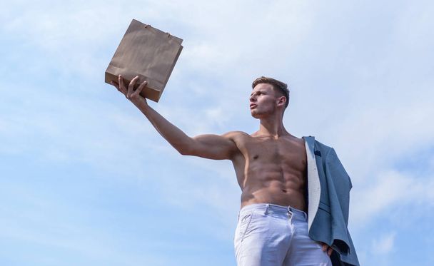 Hot sales and discount. Guy attractive macho carry shopping bag made out of brown paper. Eco package. Sales season. Shop assistant concept. Man muscular athlete hold shopping bag sky background - 写真・画像