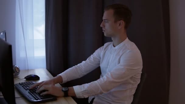 Biznesmen dorywczo lub frelancer pracy w domu, siedząc przy biurku, wpisując na klawiaturze, patrząc na ekran komputera. Szare zasłony na tle. Widok z boku - Materiał filmowy, wideo