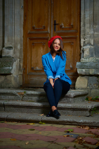 Young woman in red beret and blue jacket is sitting on a concrete porch near wooden doors. - Фото, изображение