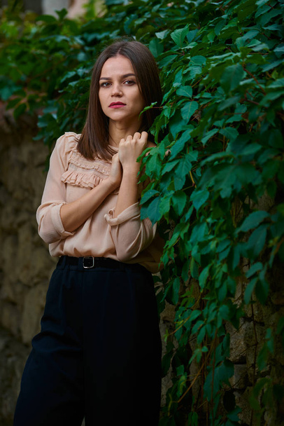 Young woman in beige blouse is posing along a stone fence covered with green leaves. - Fotó, kép