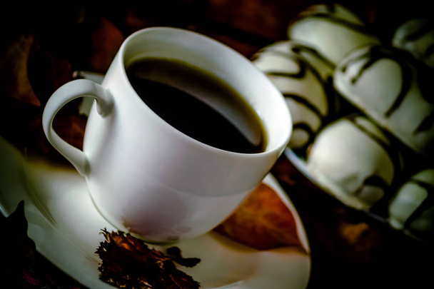 witte kop en schotel met thee of koffie is op de tafel, herfst oranje bladeren, ernaast een bord met witte koekjes met chocolade strepen, zeer donkere foto, zwart, oblique schieten, duisternis, - Foto, afbeelding