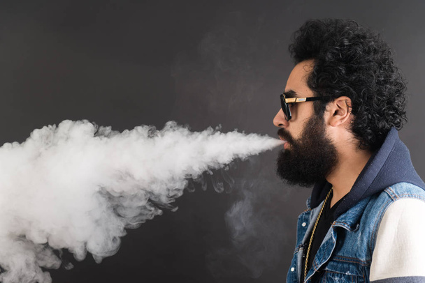 Mladý muž vapingu, studio zastřelen. Vousatý chlap s brýle zvířený oblak kouře na černém pozadí. Základní informace o kouření a páry bez nikotinu. - Fotografie, Obrázek