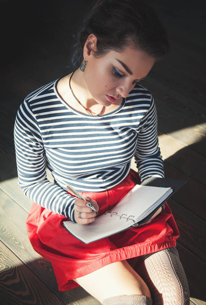 Cozy photo of young woman writing in notebook sitting on the floor in sunlight - Fotografie, Obrázek