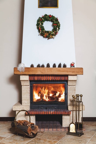 modern fireplace decorated with a Christmas wreath and a Santa Claus statue. Before the fireplace there is a forged drovnitsa and a set of brass tool - Fotografie, Obrázek