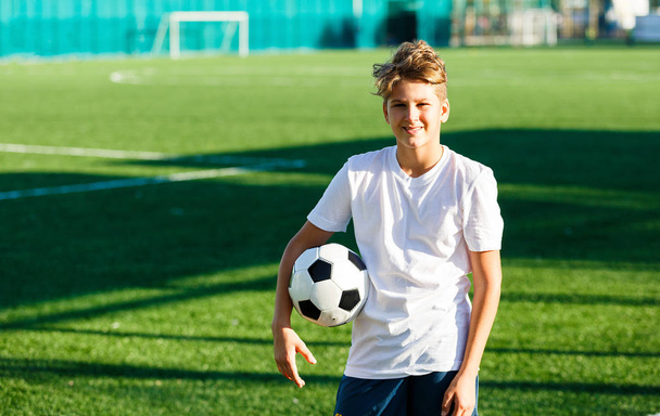 cute young boy in white blue sportswear holds classical black and white football ball on the green grass of the stadium field. Soccer game, training, hobby concept. with copy space - Photo, Image