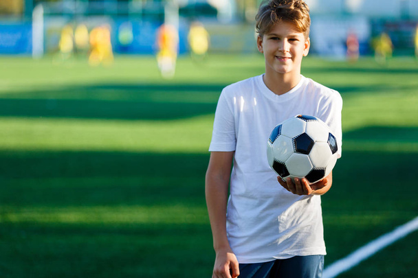 cute young boy in white blue sportswear holds classical black and white football ball on the green grass of the stadium field. Soccer game, training, hobby concept. with copy space - Foto, imagen