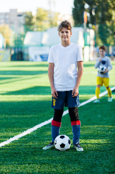 Netter kleiner Junge in weiß-blauer Sportbekleidung steht neben klassischem schwarz-weißem Fußball auf dem Stadionfeld. Fußballspiel, Training, Hobbykonzept.  - Foto, Bild