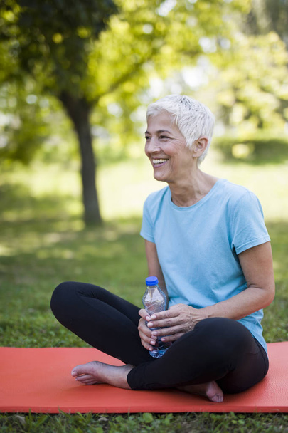 Portrait of smiling senior woman relaxing after exercising while sitting on grass - Photo, Image