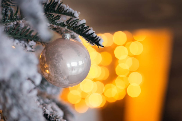 Nahaufnahme einer weißen Glaskugel auf einem Ast eines Weihnachtsbaums vor dem Hintergrund verschwommener Lichter aus der Girlande - Foto, Bild