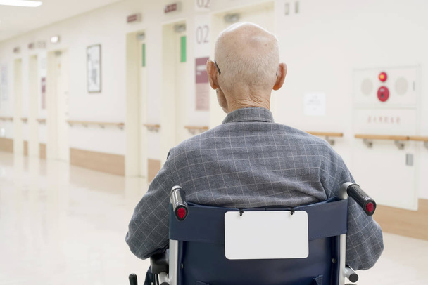 Πίσω όψη του γέροντα να κάθεται σε αναπηρικό καροτσάκι στο διάδρομο του νοσοκομείου - Φωτογραφία, εικόνα