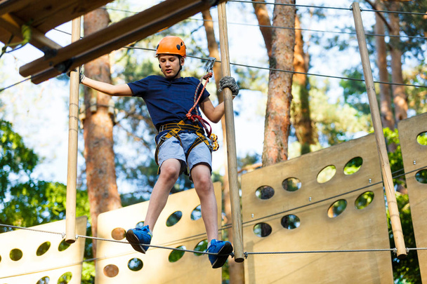 Child in forest adventure park. Kid in orange helmet  and blue t shirt climbs on high rope trail. Agility skills and climbing outdoor amusement center for children. young boy plays outdoors. - Photo, Image