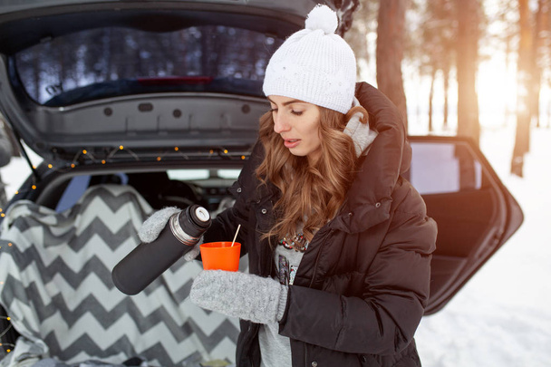 Young woman in woolen hat and black jacket stand neartrunk of the car and holds a cup of hot tea in her hands - Photo, image