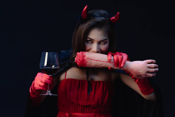 Studioaufnahme der schönen asiatischen Frau in rotem Tuch wie eine Hexe oder ein Teufel, die Horn auf dem Kopf trägt, Weinglas hält und so tut, als ob sie Menschen isst. Konzept für Halloween-Festival lustiges Cosplay. - Foto, Bild