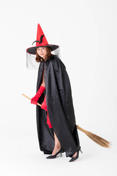 Frau in rotem Kleid Kostüm für die berühmte Hexe hält Besenstiel auf weißem Hintergrund mit Kopierraum. Konzept für lustige Aktivitäten beim Halloween-Fest. - Foto, Bild