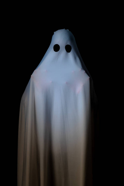Кто-то, покрытый белой тканью с большими черными глазами на черном фоне, ночью выглядит как призрак. Концепция забавной игры на Хэллоуинском фестивале
. - Фото, изображение