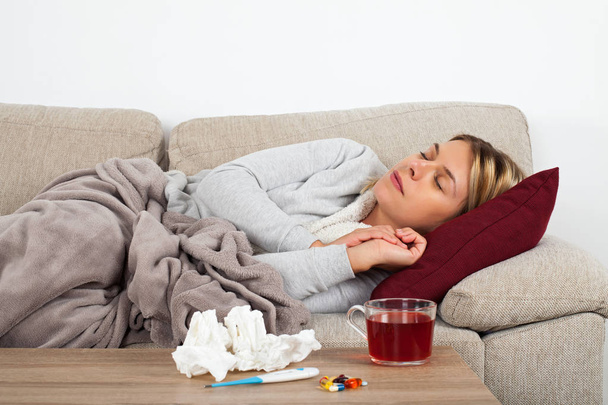 Sairas nainen makaa sohvalla käärittynä lämpimään peittoon - lääkitys, lämpömittari ja tee pöydällä
 - Valokuva, kuva