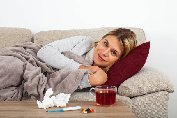 Mulher doente deitada no sofá envolta em um cobertor quente medicação, termômetro e chá na mesa
 - Foto, Imagem