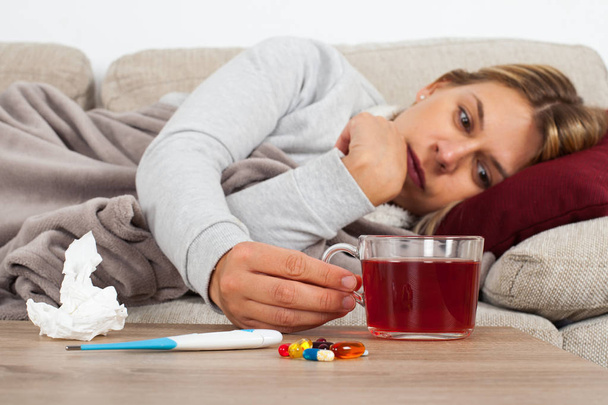 Больная женщина лежит на диване, завернутая в теплое одеяло - лекарства, термометр и чай на столе
 - Фото, изображение