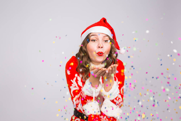 Concepto de vacaciones, Navidad y personas - Mujer hermosa y joven en traje de Santa Claus sobre fondo blanco soplando en confeti
 - Foto, imagen