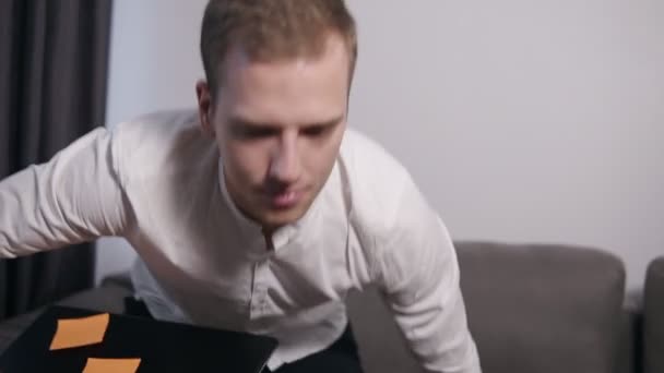 Young man in white shirt sits in the grey sofa at home and starting work on his laptop holding it on his legs. White colored wall on the background - Video