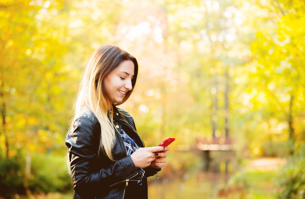 Young white girl using a mobile phone in a park with yellow trees on background. Autumn season time - Photo, Image