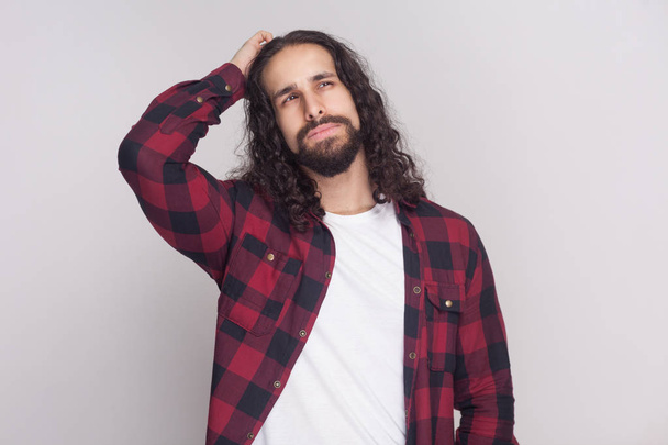 confused handsome man with beard and black long curly hair in checkered red shirt thinking and looking away with thoughtful face on grey background  - Fotografie, Obrázek
