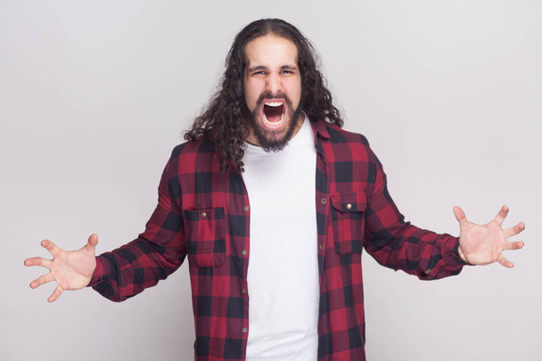 Angry handsome man with beard and black long curly hair in checkered red shirt looking at camera and shouting with raised arms on grey background. - Φωτογραφία, εικόνα