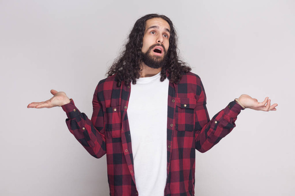 Portrait of confused man with beard and black long curly hair in checkered red shirt standing with raised arms on grey background - Foto, afbeelding