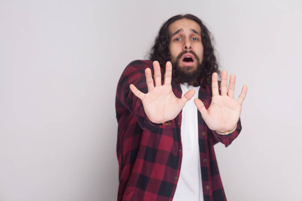 Afraid man with beard and black long curly hair in checkered red shirt looking at camera with blocking hands on grey background - Фото, изображение